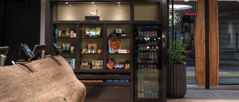 Een moderne hotel lobby met een grote houten sculptuur, een minibar gevuld met snacks en drankjes, en een glazen deur naar buiten.