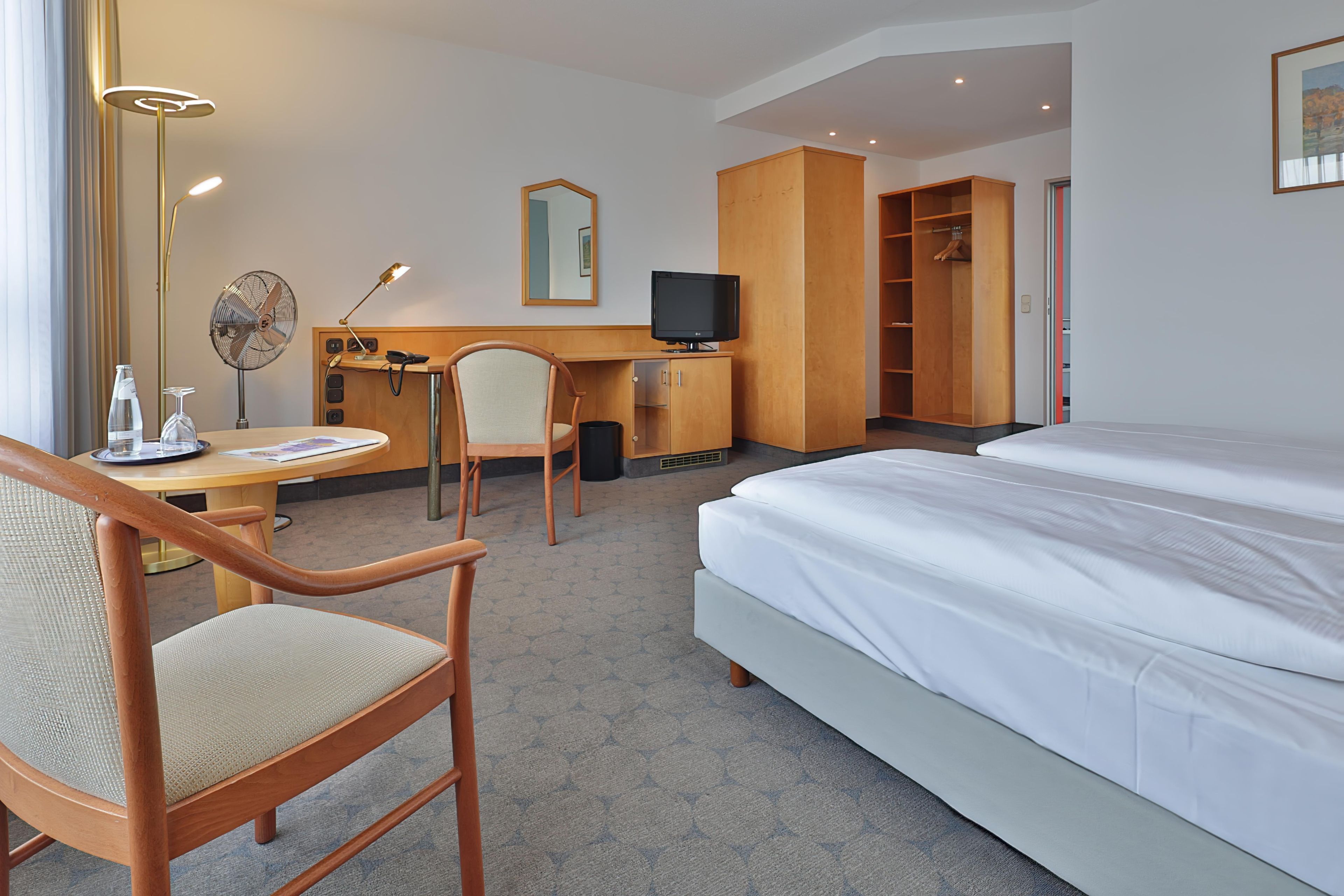 A hotel room with a bed, a desk with a chair, a TV, a wardrobe, and a mirror.