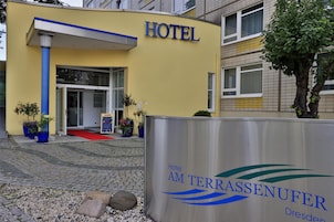A hotel entrance with a yellow facade, a blue sign, and a signboard for Hotel Am Terrassenufer.