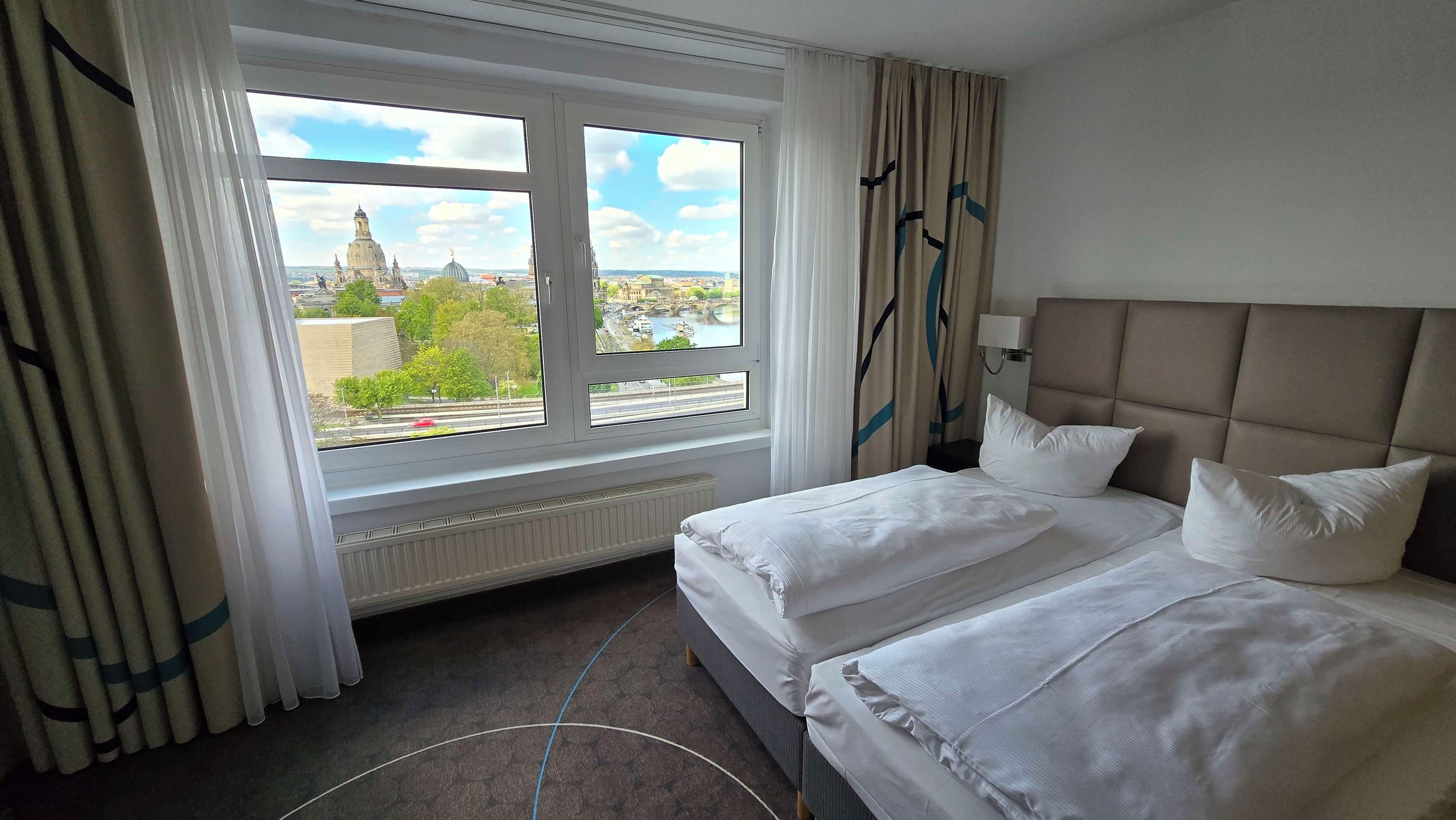 A hotel room with a bed, a window with curtains, a radiator, and a view of a cityscape.