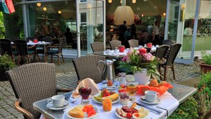 Un espace extérieur aménagé pour prendre les repas, avec des chaises en osier, une table dressée avec des aliments pour le petit-déjeuner et un journal.