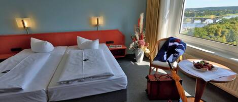 A hotel room with two beds, a small table with a fruit bowl, a chair, and a view of a river and bridge.