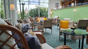 Un espace salon accueillant, avec des meubles en osier, une table en verre et une vue sur la verdure et un bâtiment en arrière-plan.