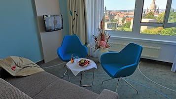 A hotel room with a view of a cityscape, two blue chairs, a table with fruit and a bottle, and a grey sofa.