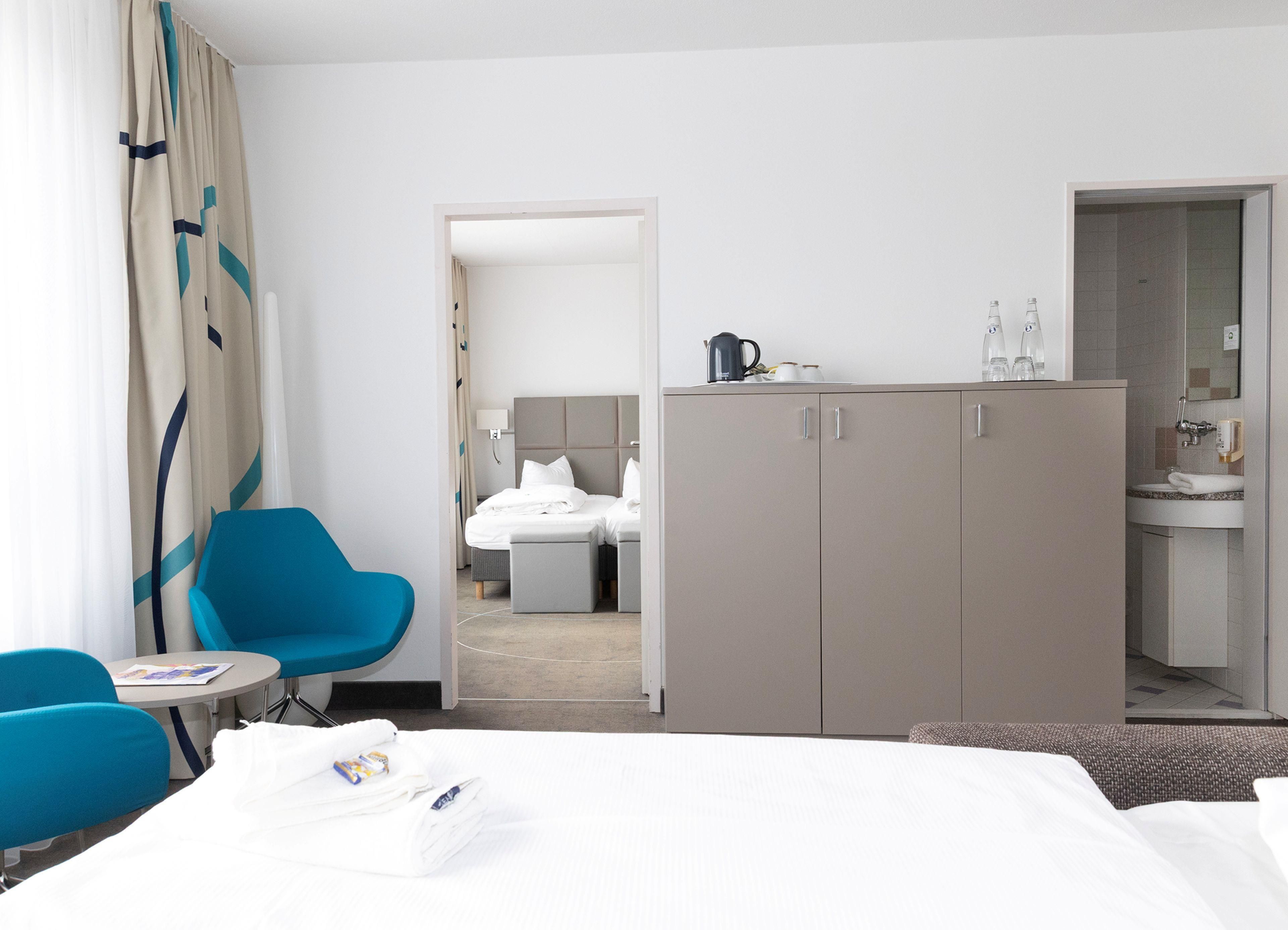 A hotel room with a bed, a blue armchair, a small table, a wardrobe, and a bathroom visible through an open door.