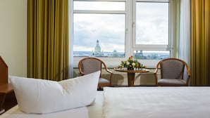 Une chambre d’hôtel avec un lit, deux chaises, une petite table agrémentée de fleurs et une vue sur le paysage urbain, visible à travers de grandes fenêtres.