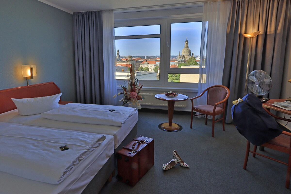 Une chambre d’hôtel avec deux lits, un bureau, une chaise, un ventilateur et une vue sur le paysage urbain.