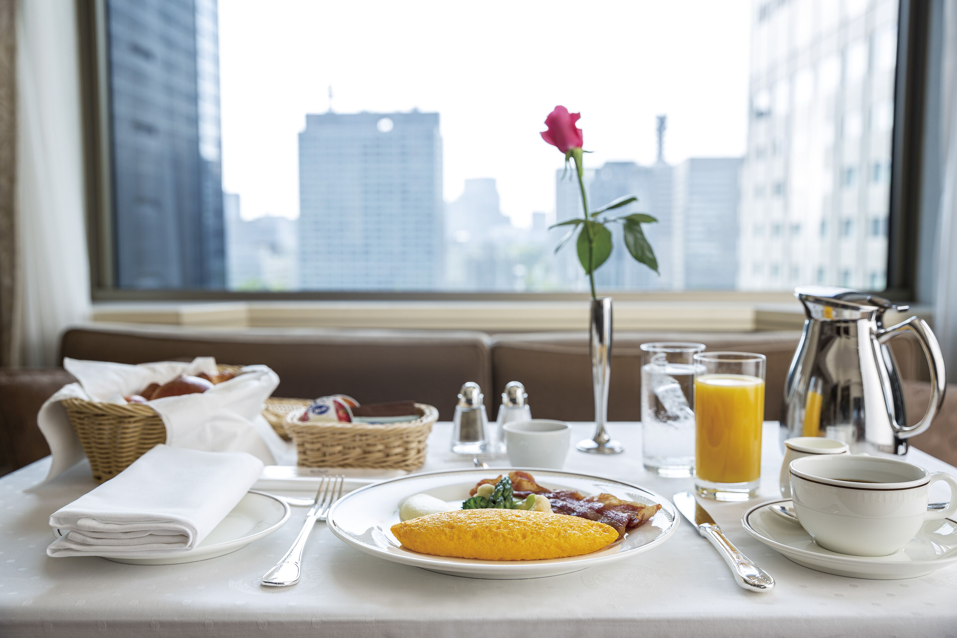 A breakfast table with a city view, including a rose, a pitcher, and a plate of food.