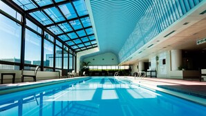 Piscina interior com teto de vidro, área de estar e vista para a paisagem urbana.