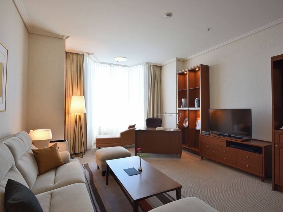 A hotel room with a sofa, coffee table, TV, and a desk with a lamp.