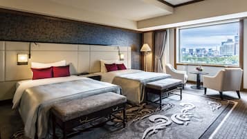 A hotel room with two beds, a small table, a chair, and a view of the city.