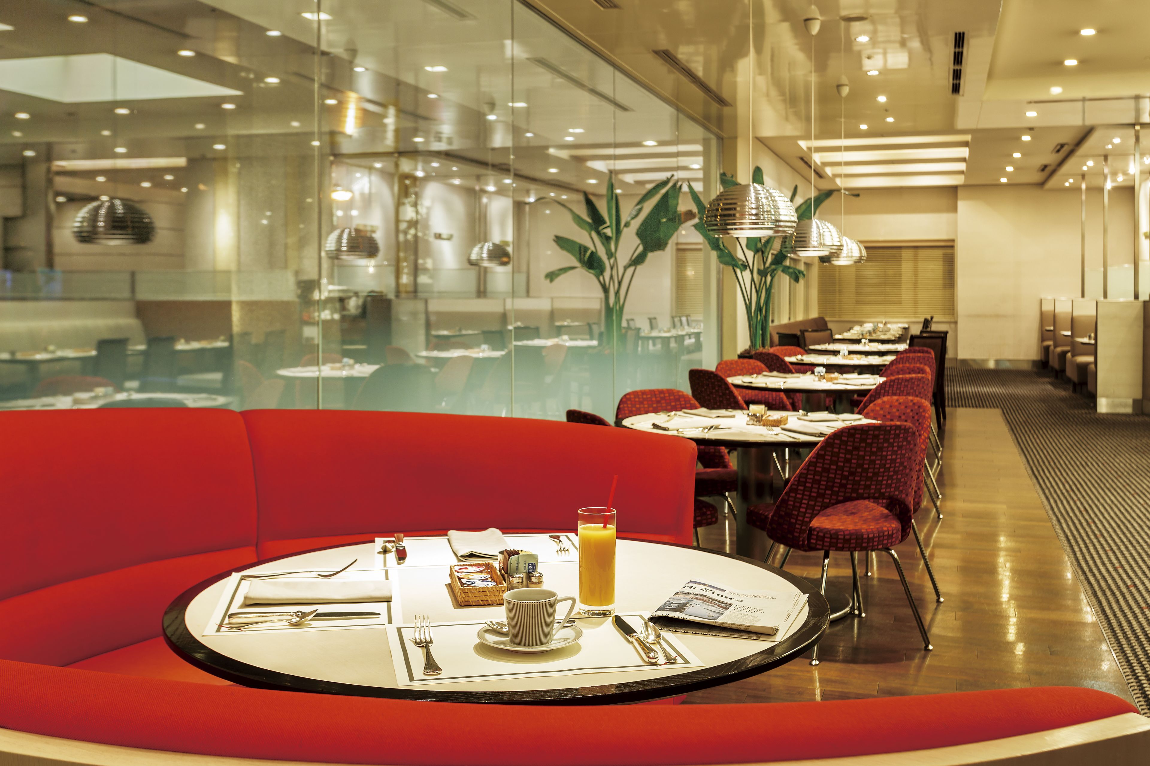A modern restaurant with red seating, a round table with a breakfast setting, and a view of the kitchen through a glass wall.