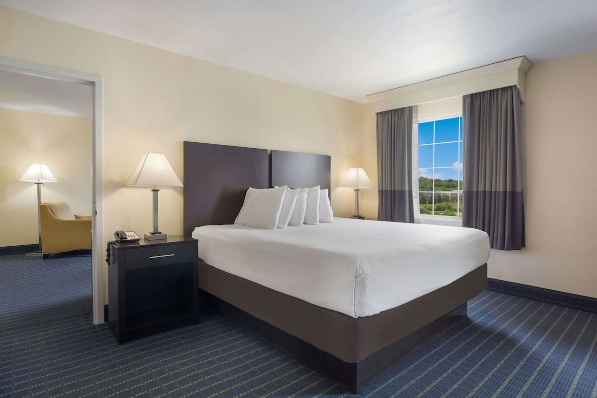 A hotel room with a large bed, two bedside tables with lamps, a chair, and a view of the outdoors through a window.