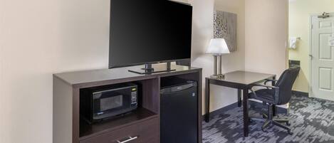 Habitación de hotel pequeña con televisión de pantalla plana, microondas, refrigerador miniatura, escritorio con silla y lámpara.
