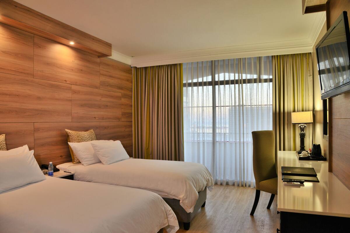 A hotel room with two beds, a desk, a chair, a TV, and a large window with curtains.