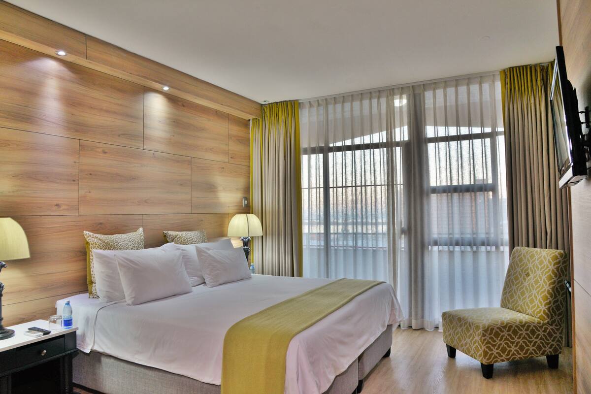 A hotel room with a wooden panel wall, a large bed with white bedding, two bedside lamps, a chair, and a TV mounted on the wall.