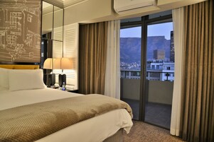 A hotel room with a large bed, a bedside table with a lamp, a window with curtains, and a view of a cityscape and mountains.