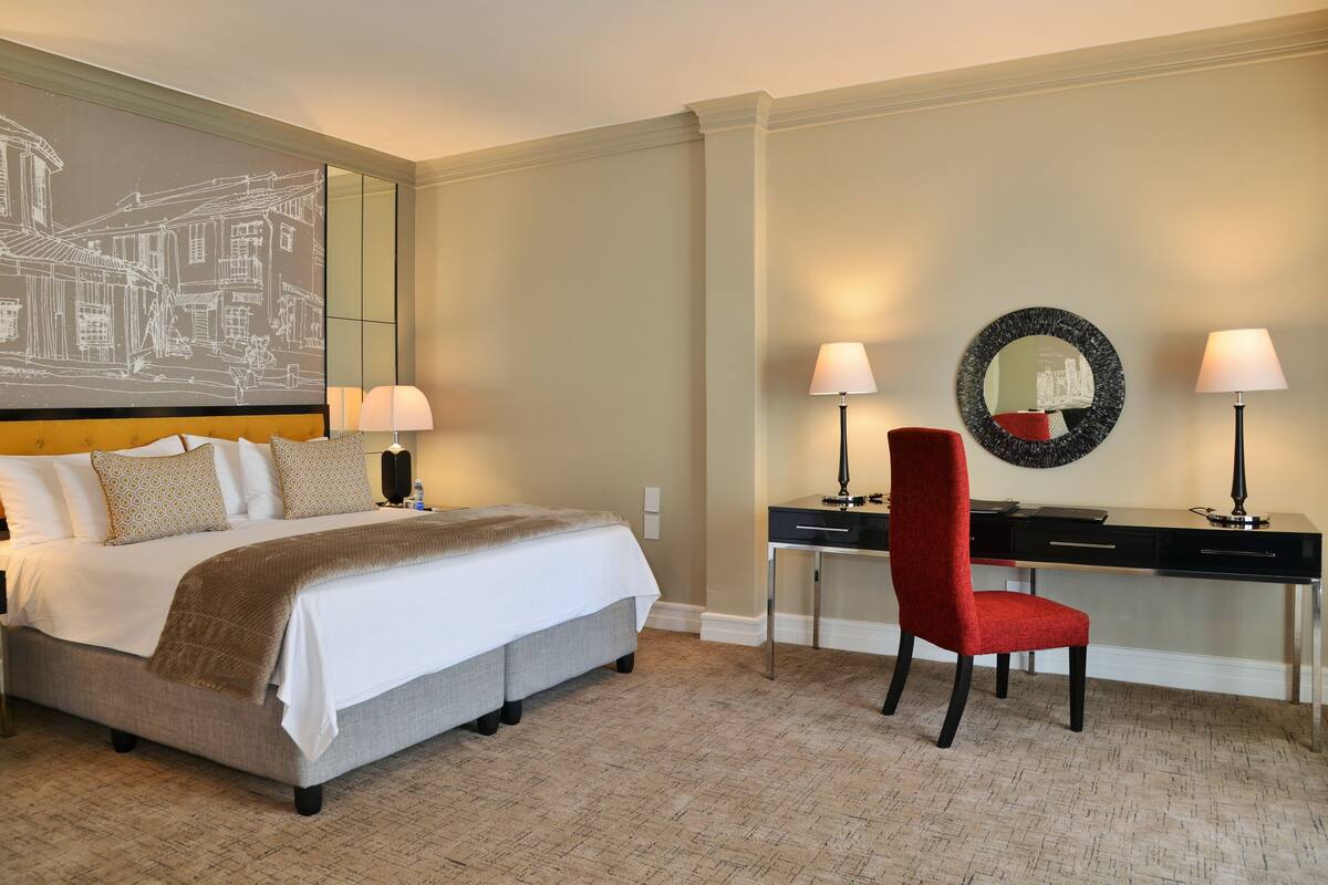 A hotel room with a large bed, a desk, a chair, and a wall-mounted mirror.