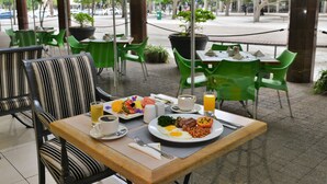 An outdoor dining area with green chairs and tables, a breakfast spread including eggs, beans, and toast, and a view of the street and trees through large windows. - Capetonian Hotel (Cape Town)