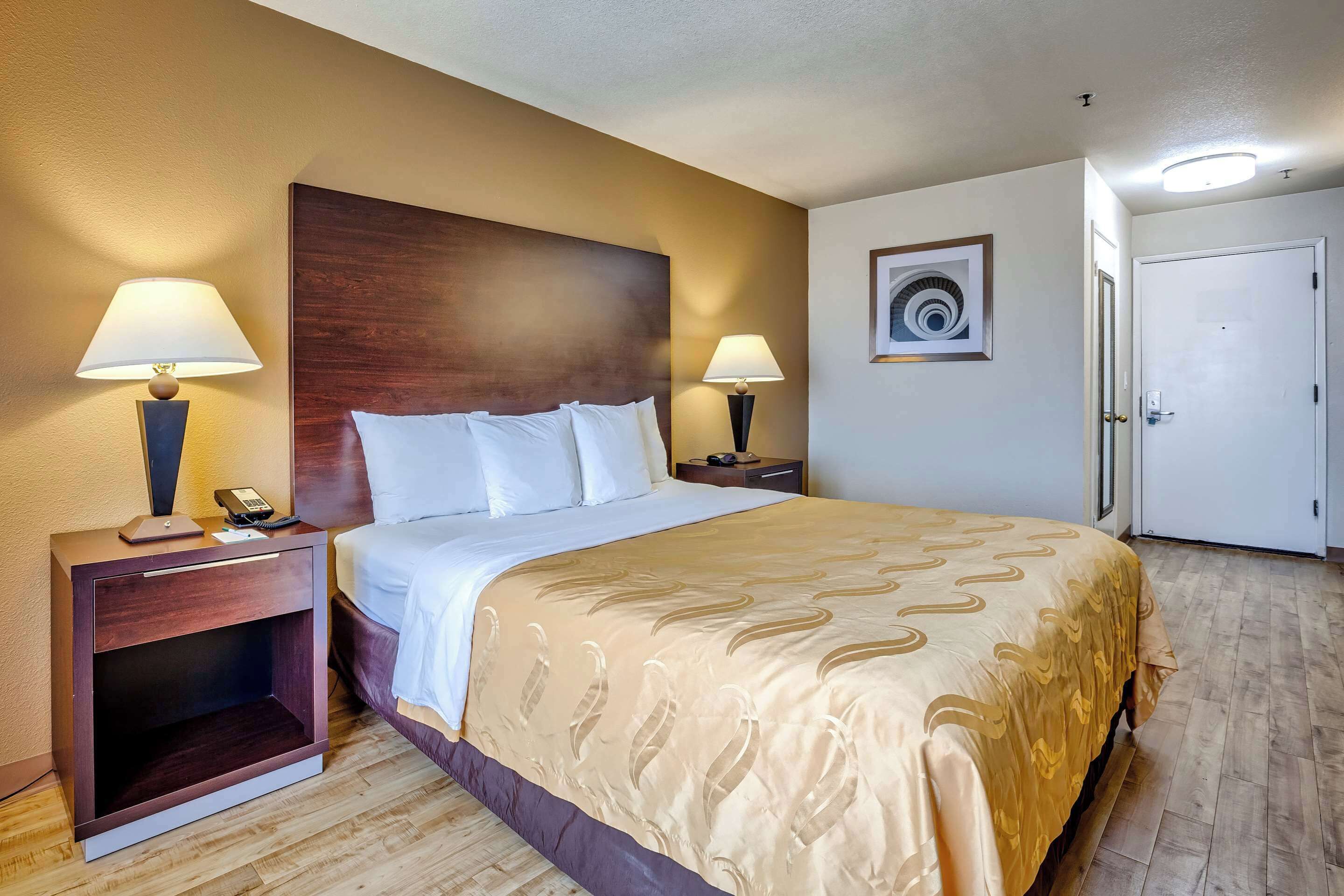 Quarto de hotel com uma cama grande, duas mesas de cabeceira com luminárias, um painel de cama de madeira, um quadro na parede e uma porta.