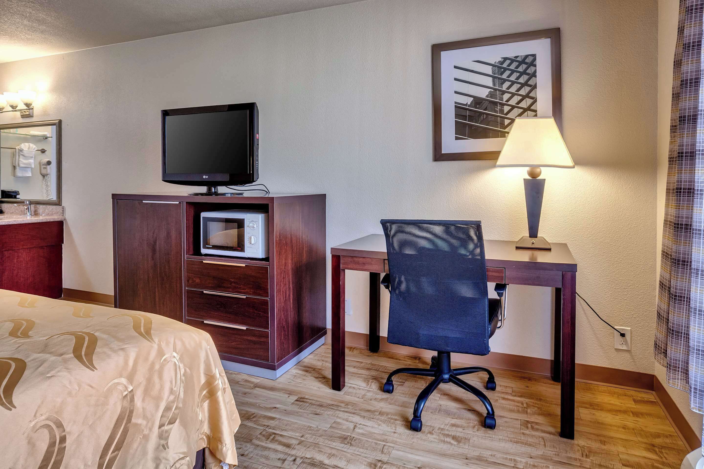 Een hotelkamer met een bed, een bureau met stoel, een televisie, een lamp en een ingelijste foto aan de muur.