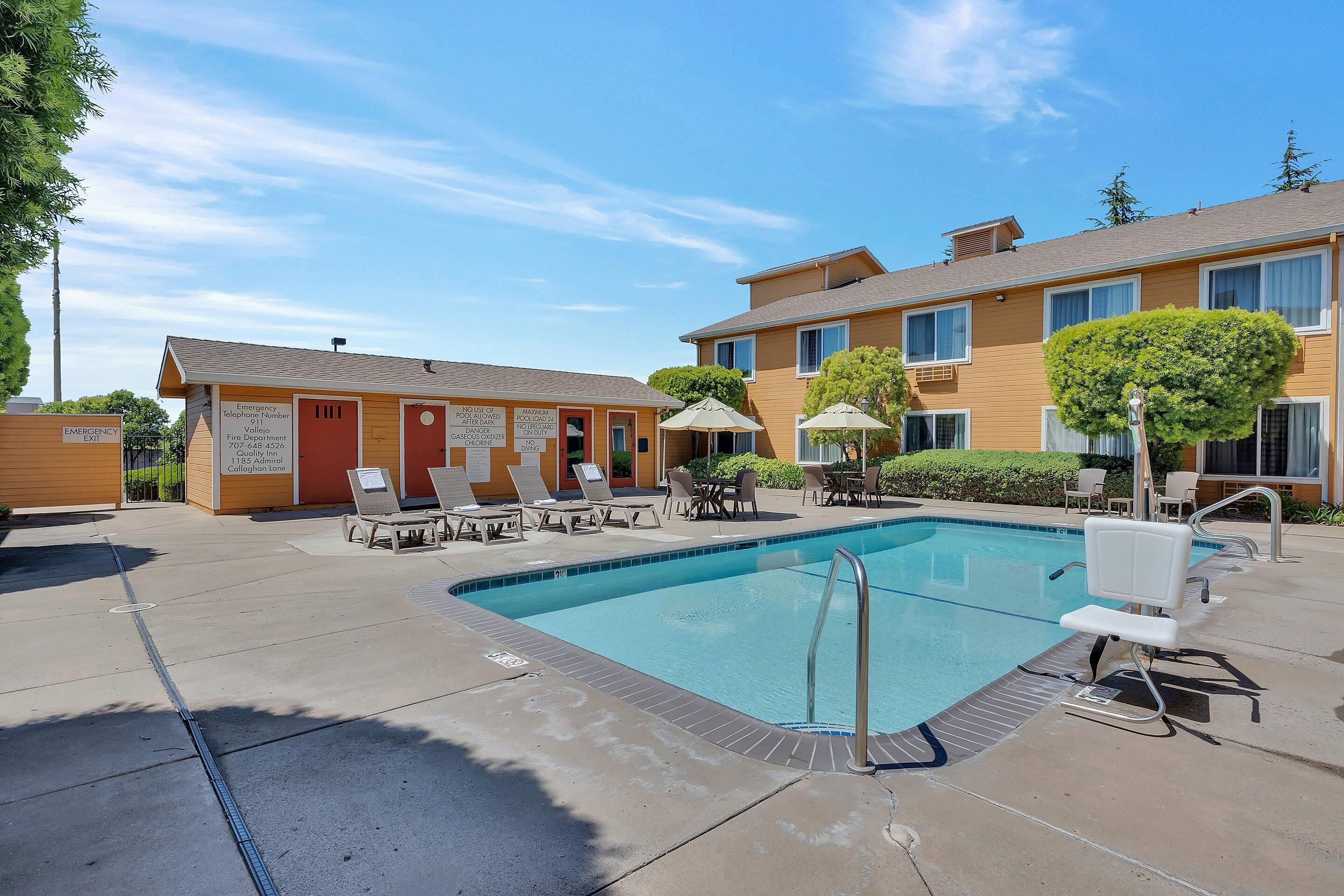 A pool area with lounge chairs, a small building labeled "Emergency," and a clear blue sky.