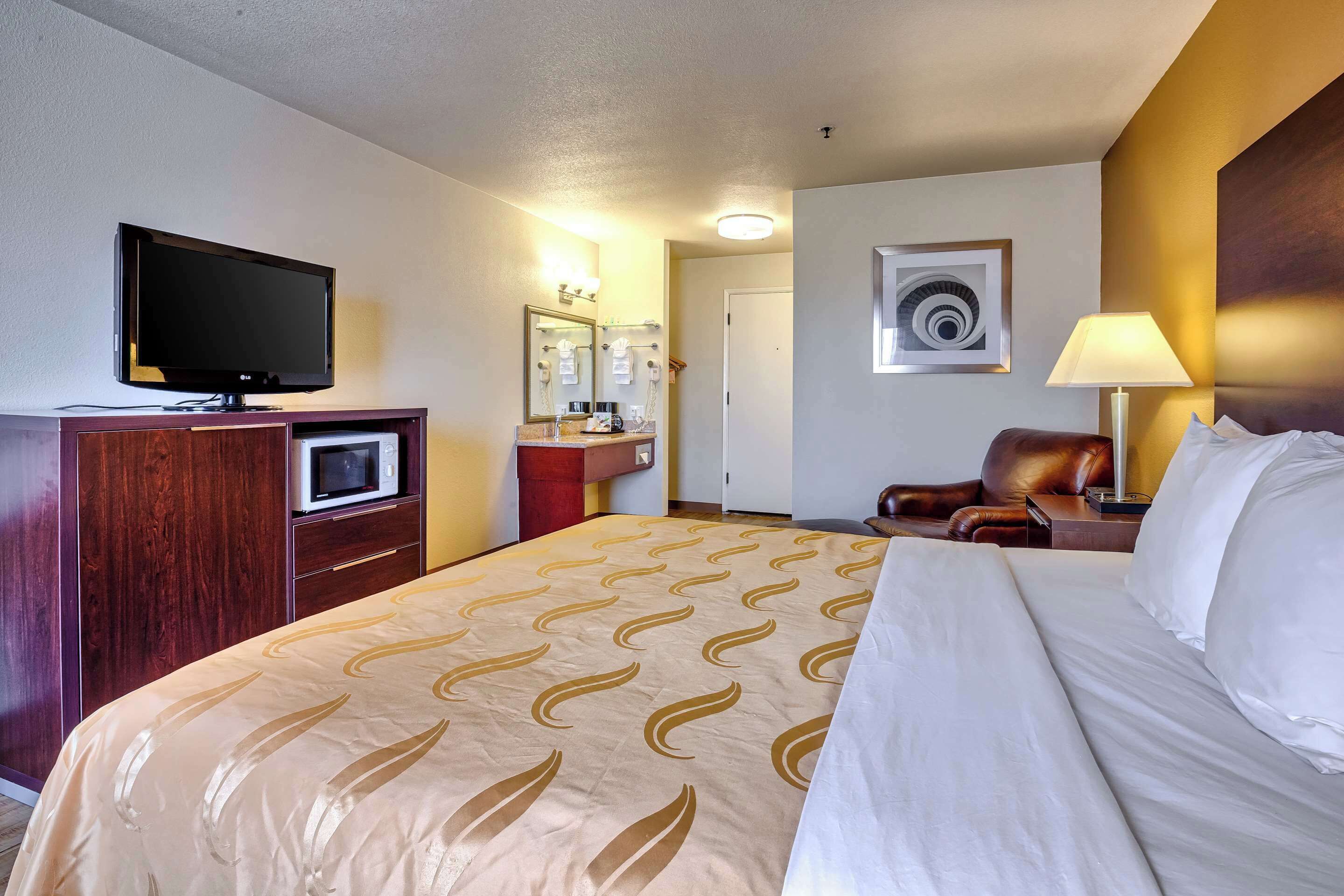 Een hotelkamer met een bed, televisie, bureau, stoel, lamp en een badkamer met spiegel en wastafel.