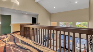 A hotel interior with a high ceiling, a wooden railing, a patterned carpet, and a view of the street through large windows.