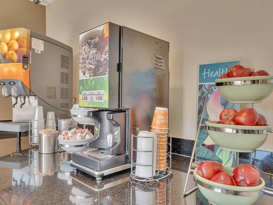 A self-service breakfast station with juice and coffee dispensers, a coffee maker, and a fruit display with apples.