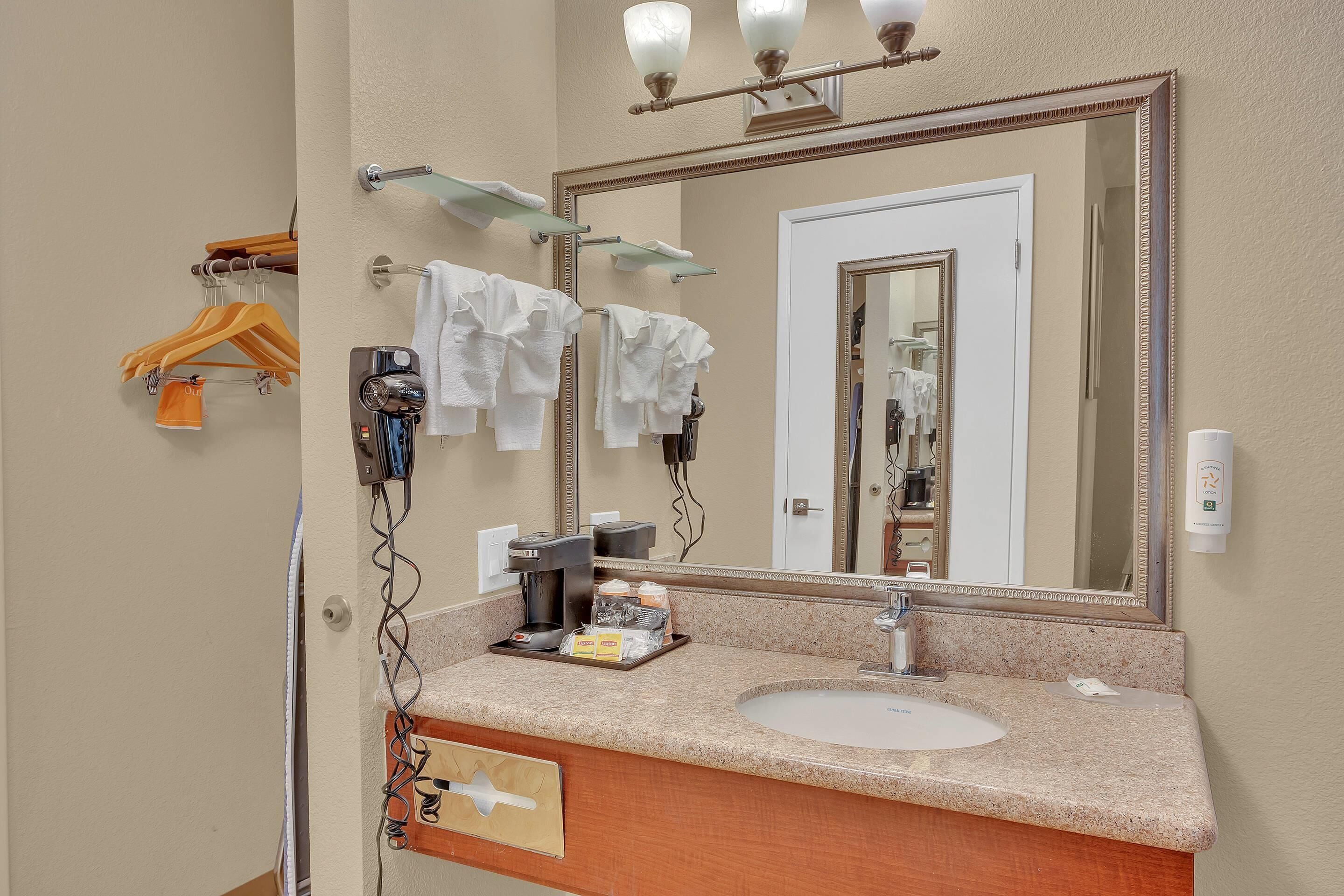 A hotel bathroom with a granite countertop, a sink, a mirror, towel racks, and a coffee machine.