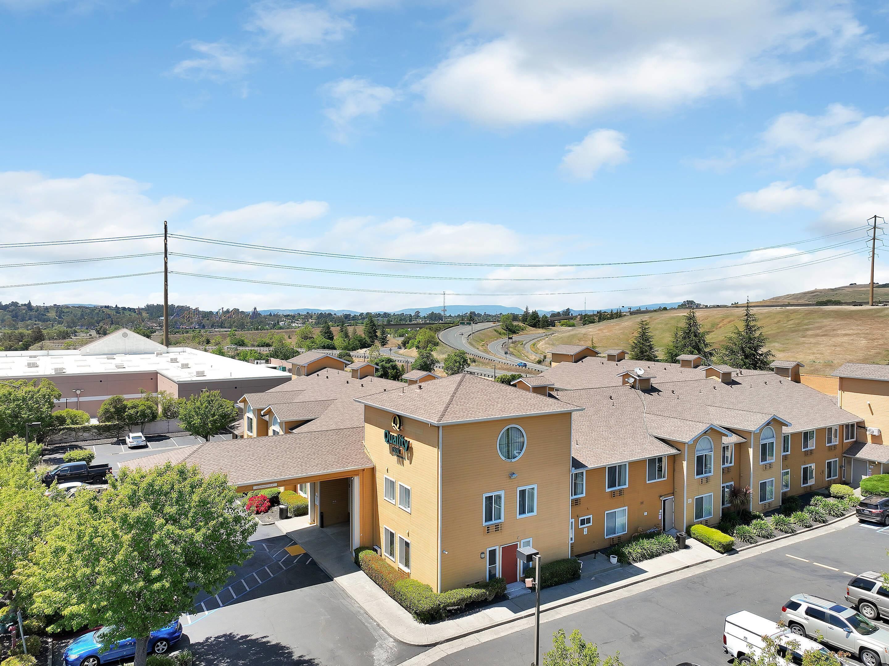 A multi-story hotel with a flat roof, a parking lot, and a view of a hilly landscape in the background.