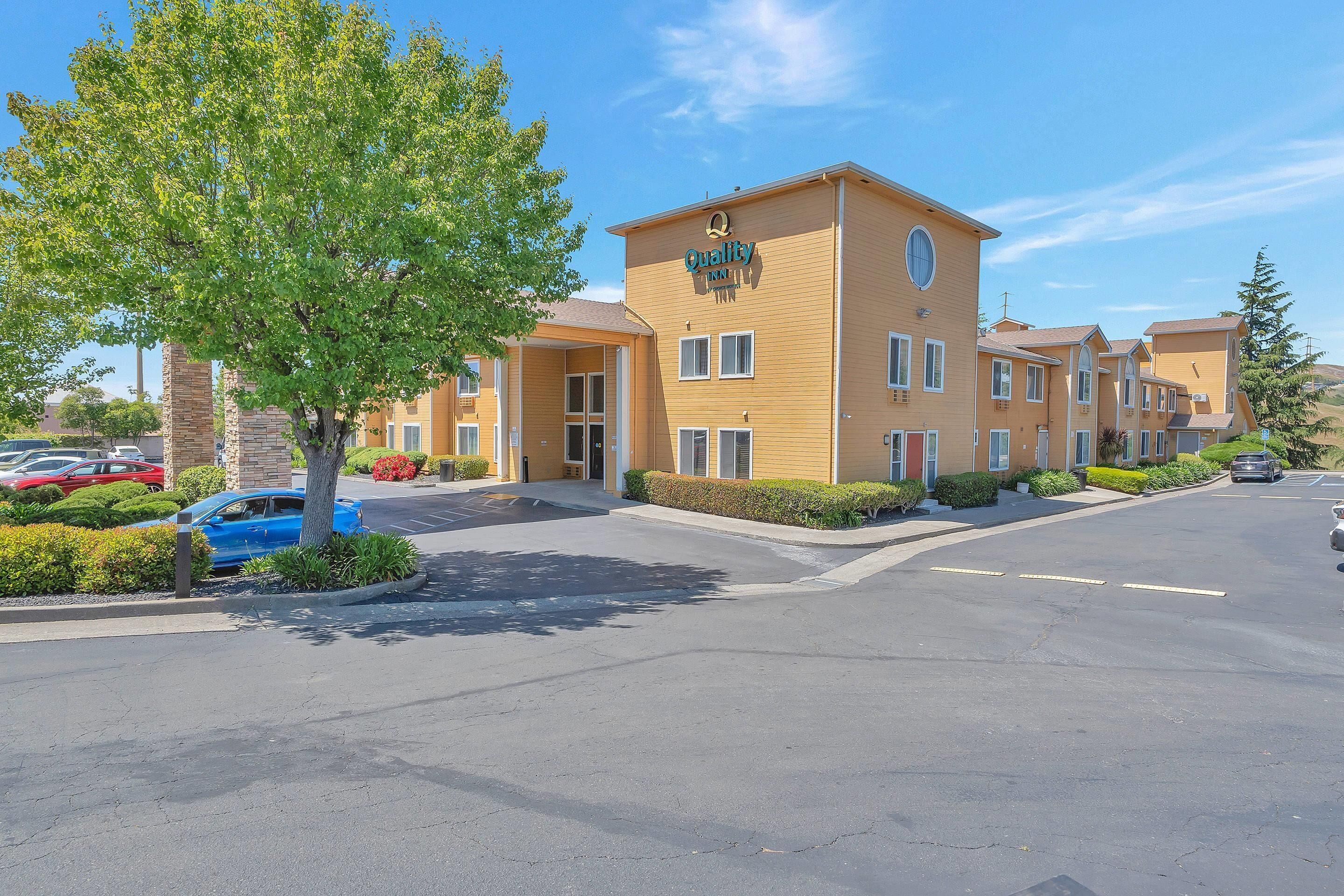 A hotel with a yellow facade, a large tree in front, and a parking lot with several cars.