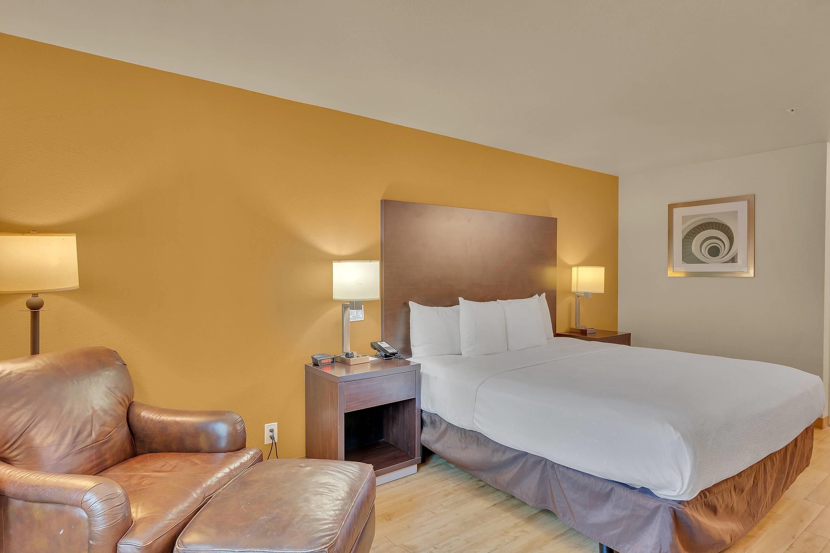 A hotel room with a bed, a leather armchair, a nightstand with lamps, and a wall-mounted artwork.