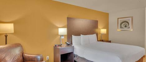 A hotel room with a bed, a leather armchair, a nightstand with lamps, and a wall-mounted artwork.