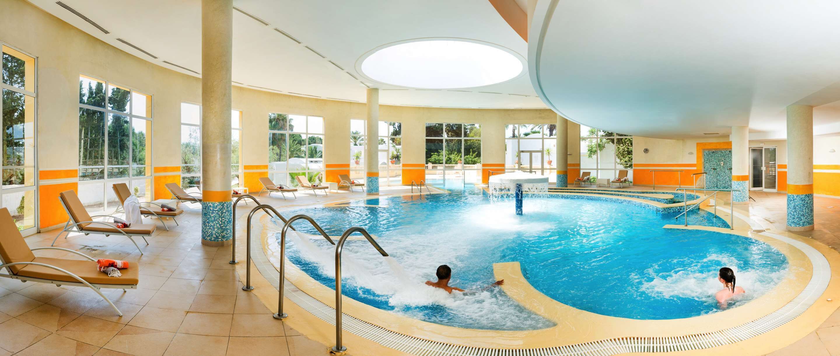 Una piscina interior con dos personas nadando, rodeada de sillones de descanso y una tumbona.