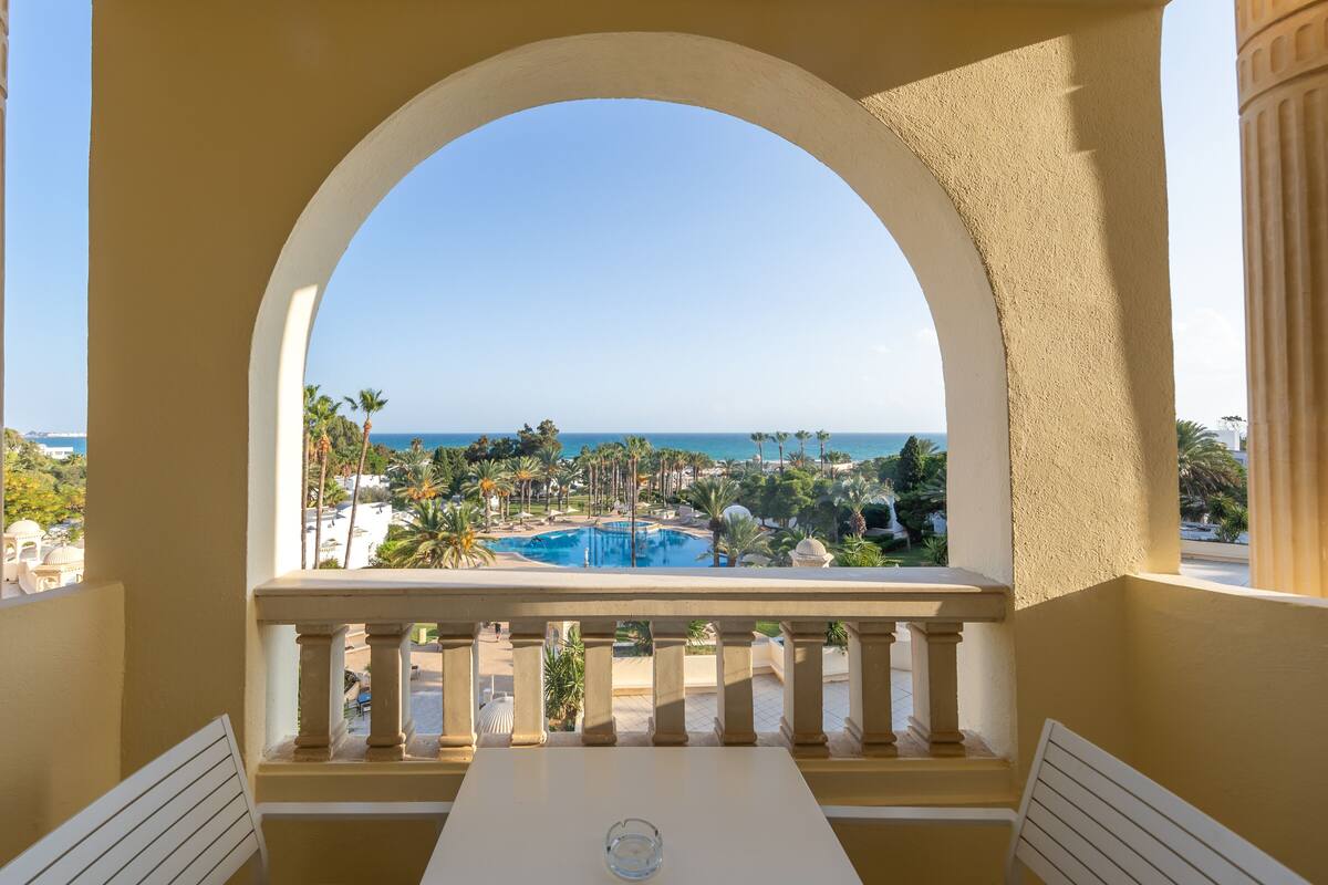 Un balcó amb vista a una piscina i palmeres, emmarcat per un arc de color beix.