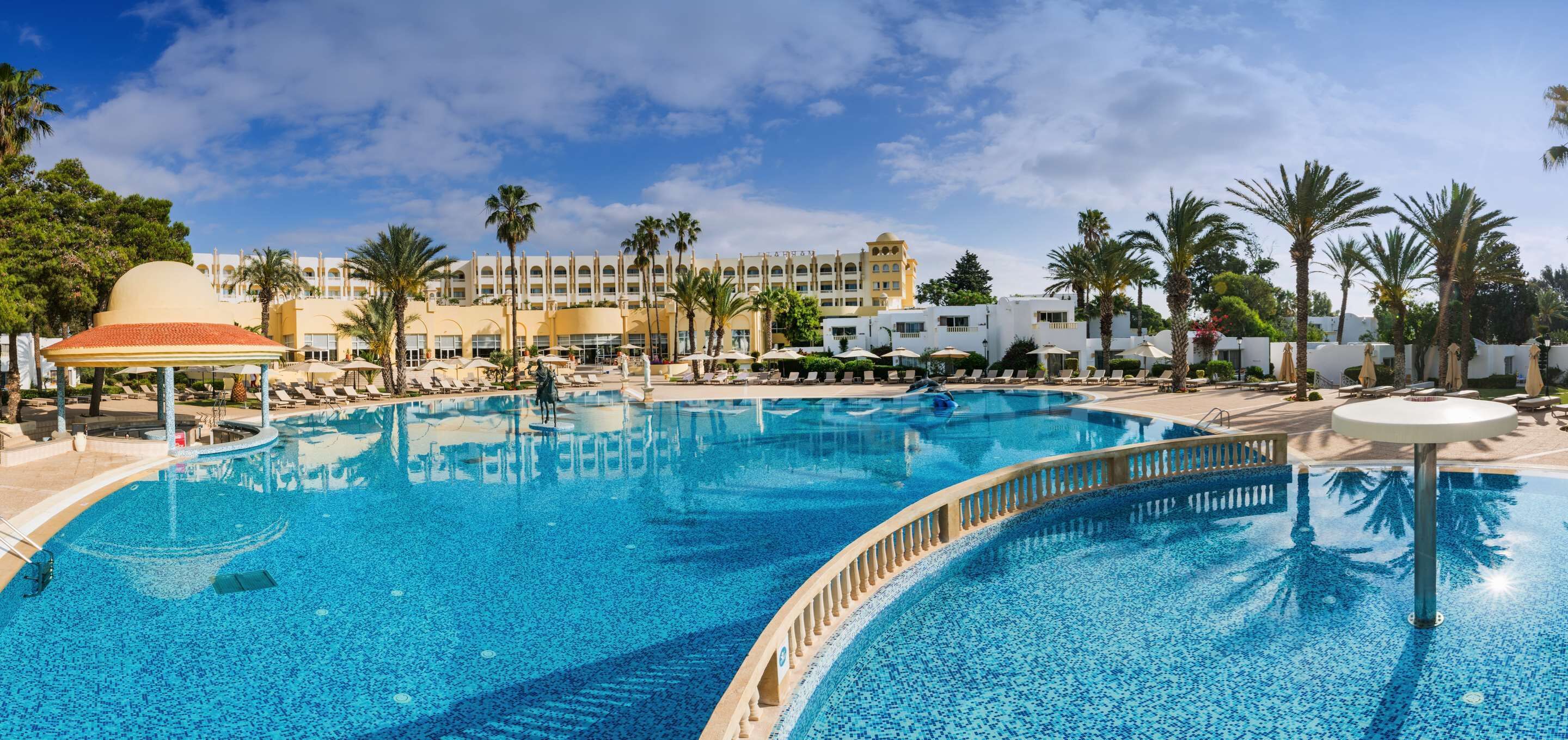 A large outdoor swimming pool with lounge chairs and umbrellas, surrounded by palm trees and a hotel building in the background.