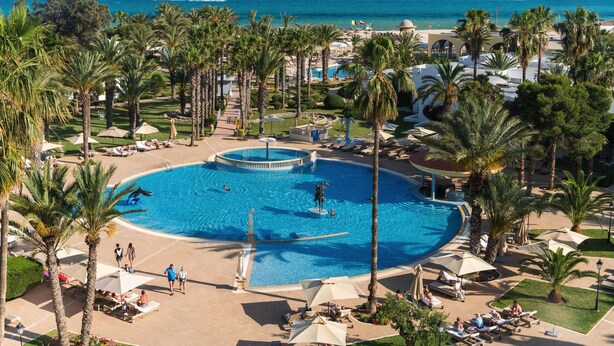 Een resort met een groot centraal zwembad, palmbomen en uitzicht op de oceaan.