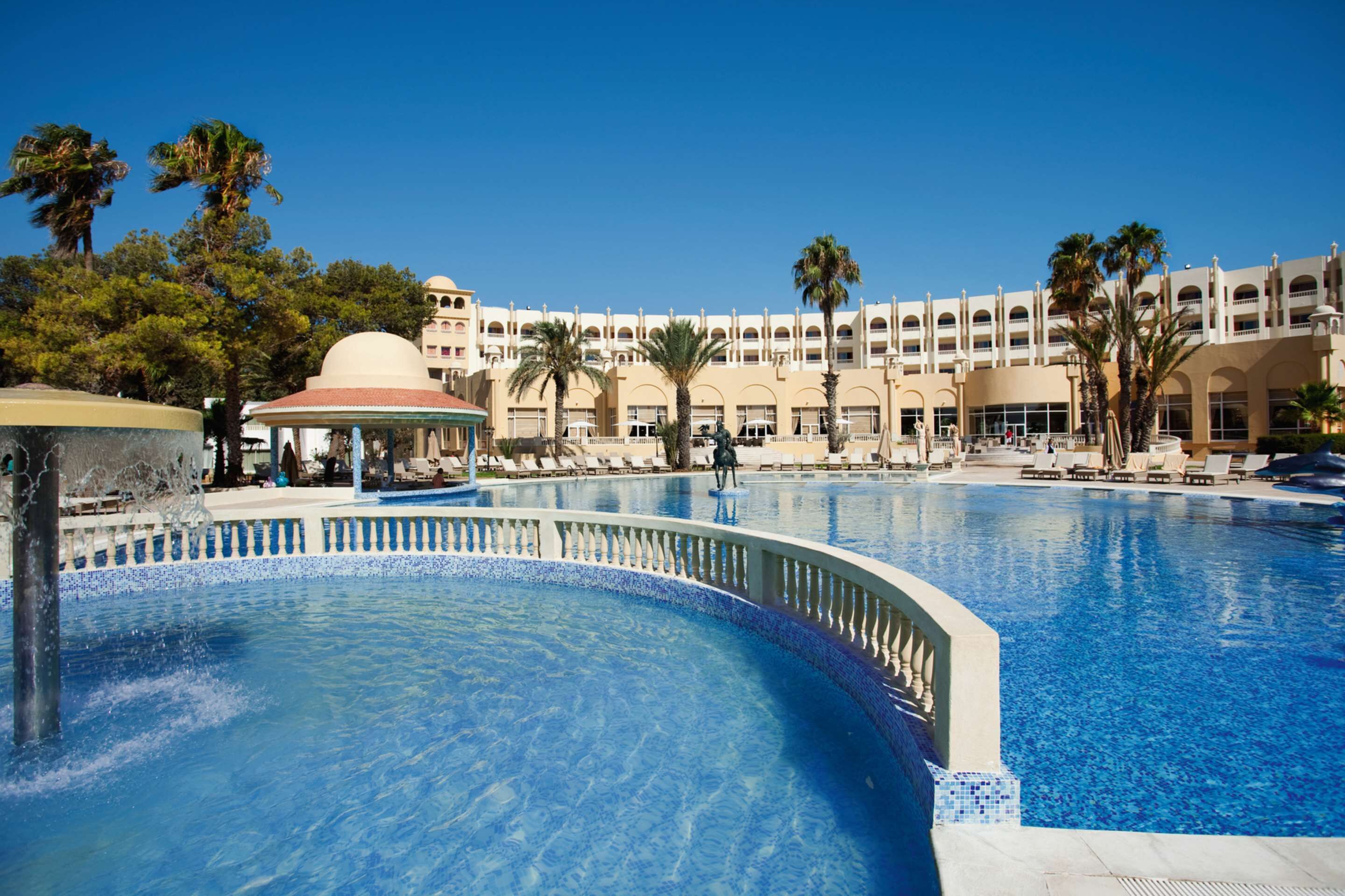 A large swimming pool with a fountain, palm trees, and a multi-story building with balconies in the background.