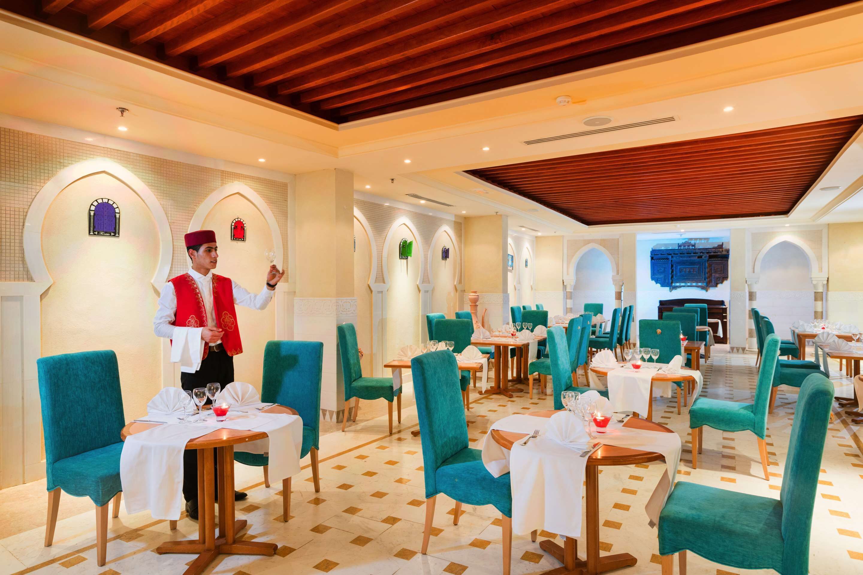 A restaurant with teal chairs and tables, a waiter in uniform, and a wooden ceiling.