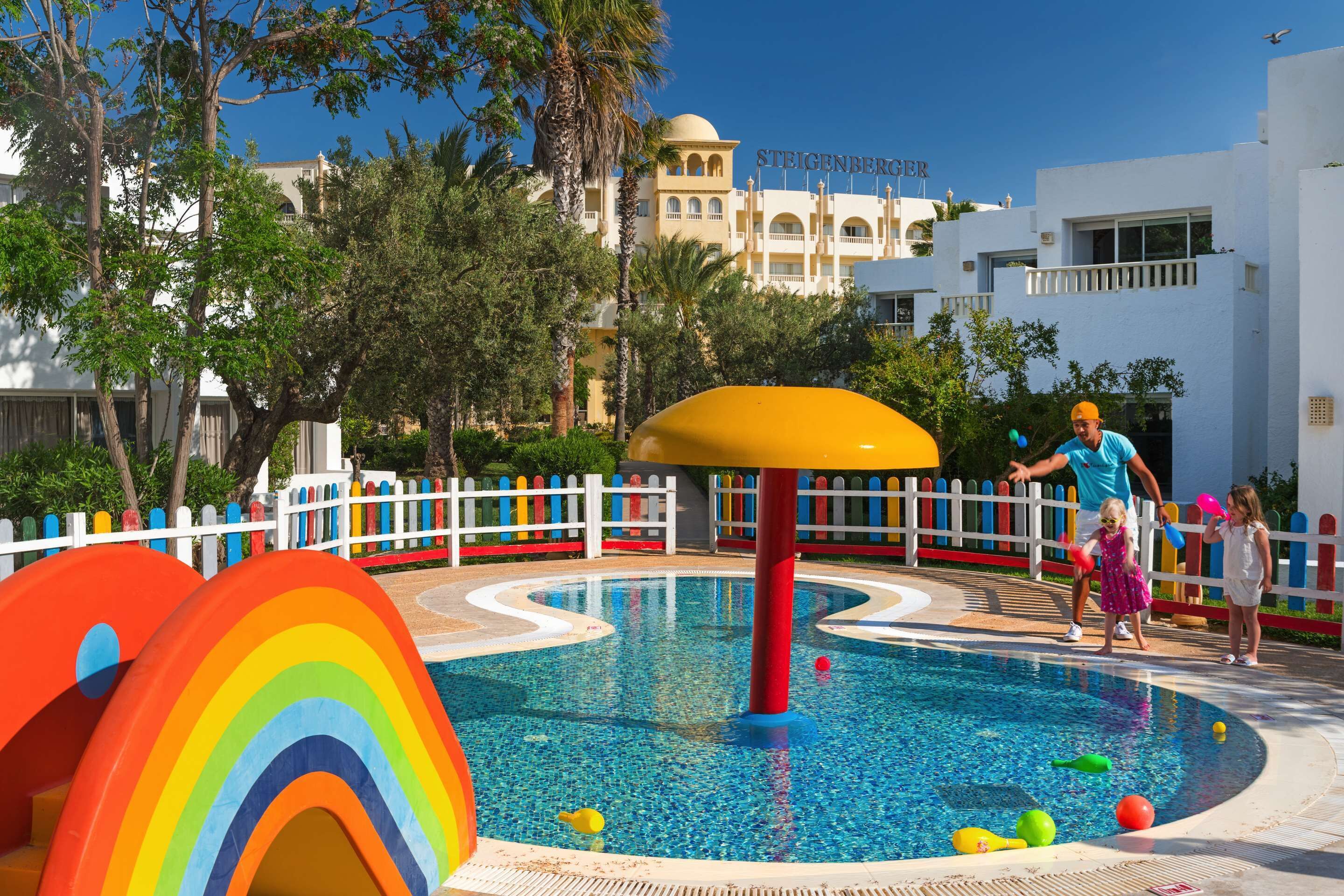 Zona de piscina con una estructura de juego colorida, una cerca blanca y un edificio con la señalización 'STEIGENBERGER' al fondo.