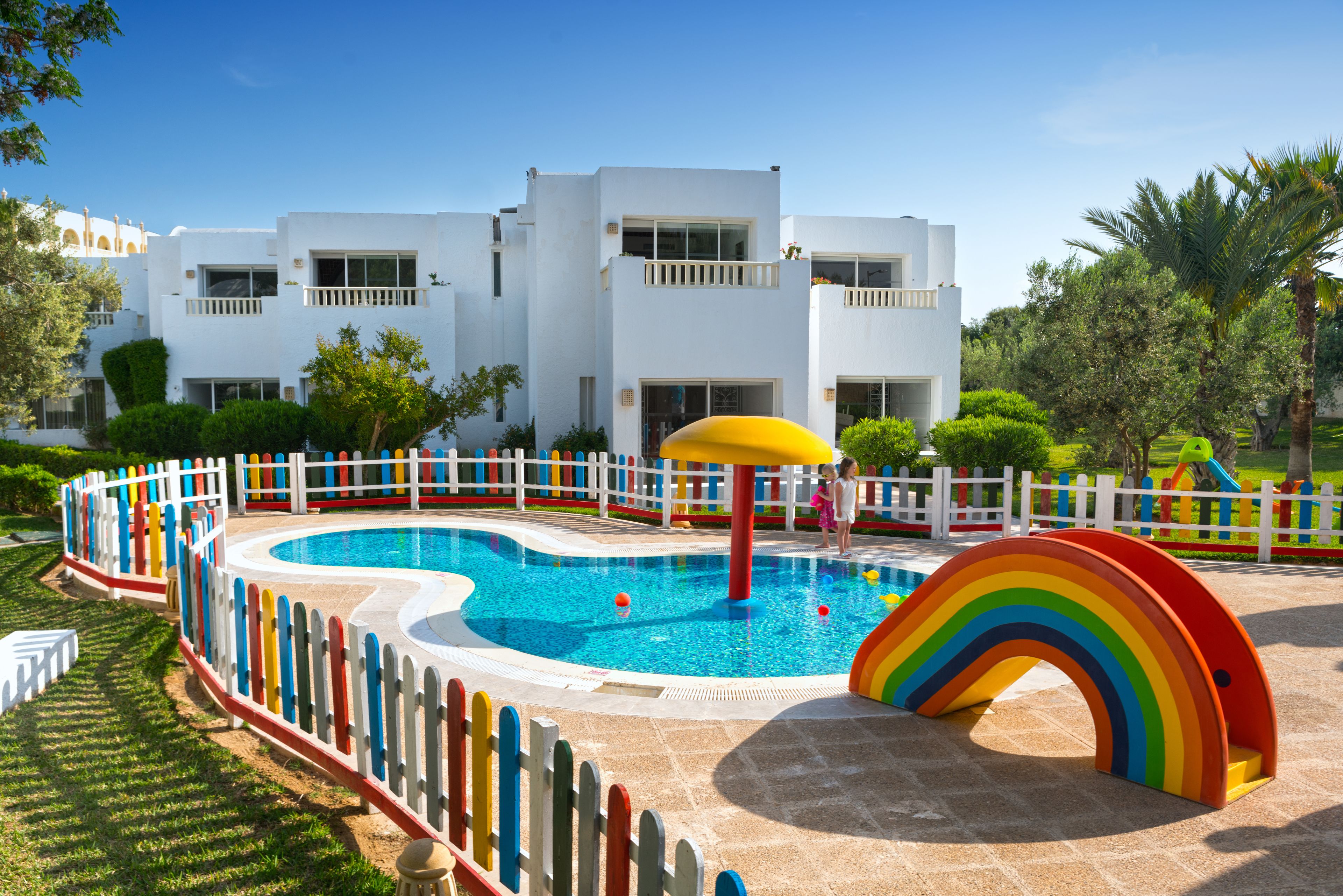 A swimming pool with a rainbow play structure and a white building in the background.
