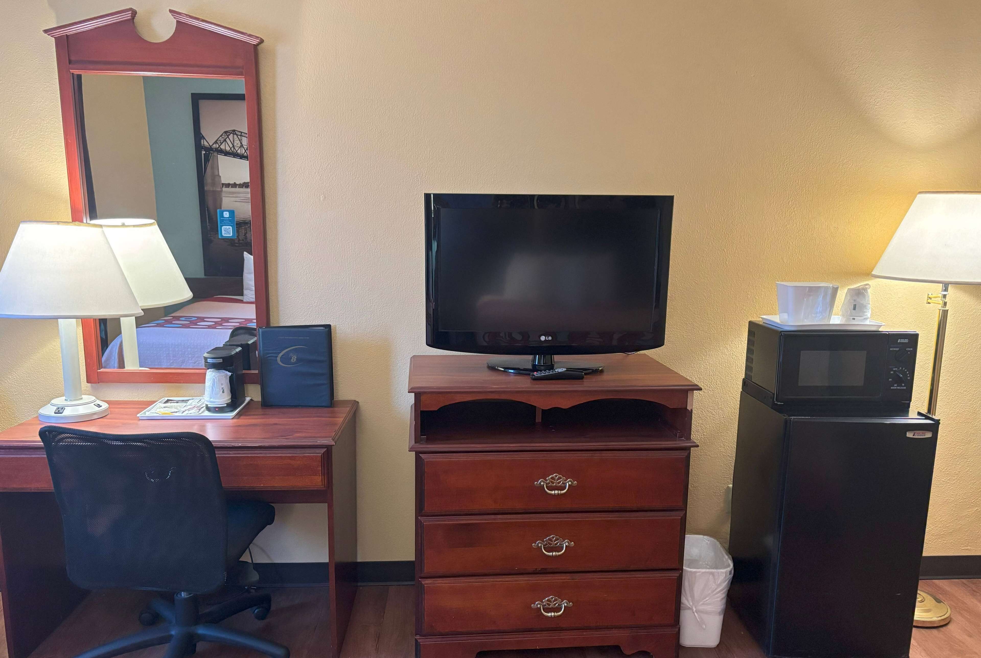 Una habitación de hotel con un escritorio de madera, una silla de oficina negra, un televisor sobre un mueble de madera, un microondas y una lámpara.