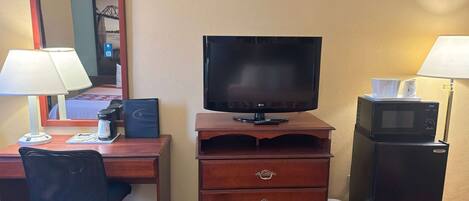 Una habitación de hotel con un escritorio de madera, una silla de oficina negra, un televisor sobre un mueble de madera, un microondas y una lámpara.