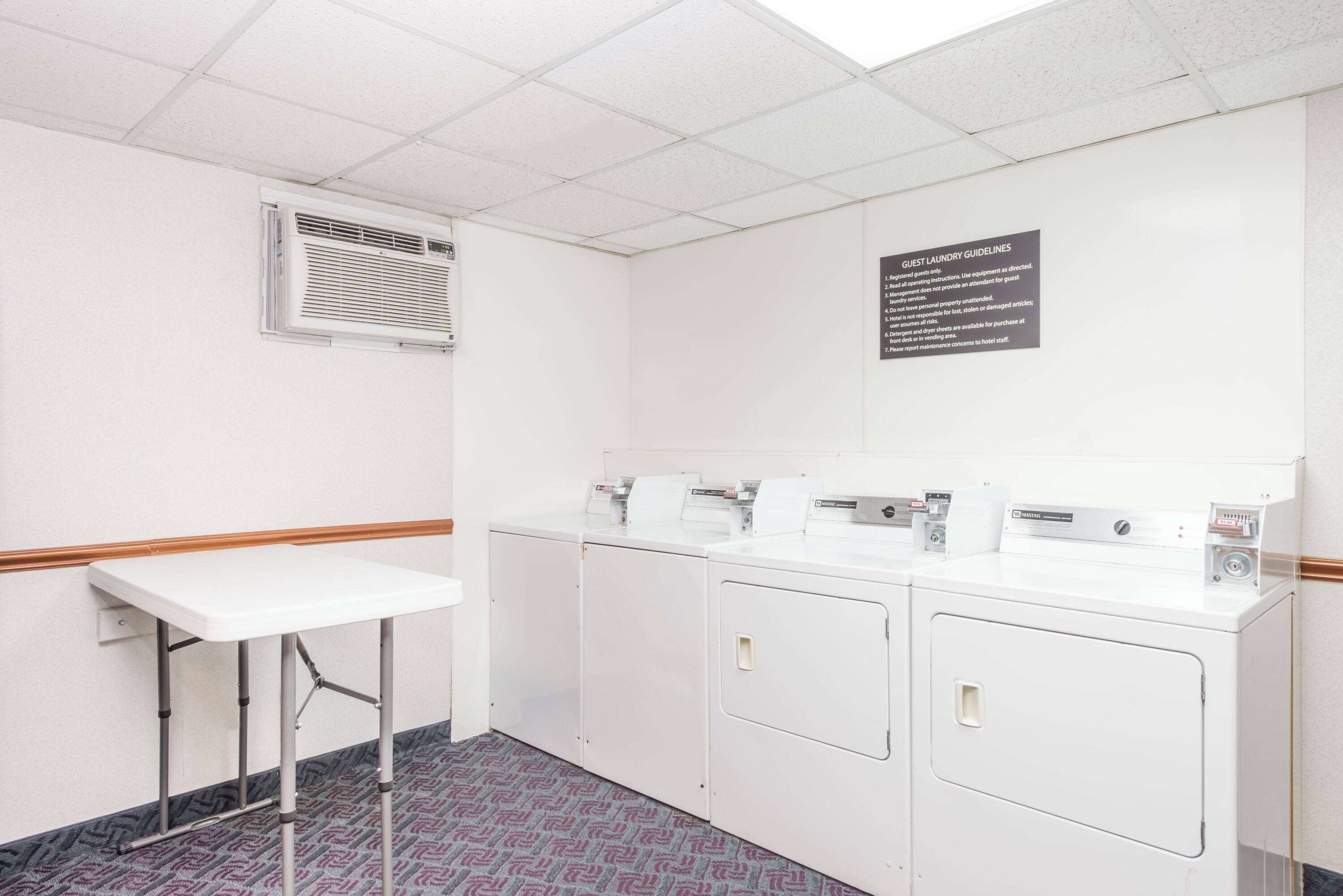 A hotel room with a white table, a washing machine, and a dryer. There is a sign on the wall with guest laundry guidelines.