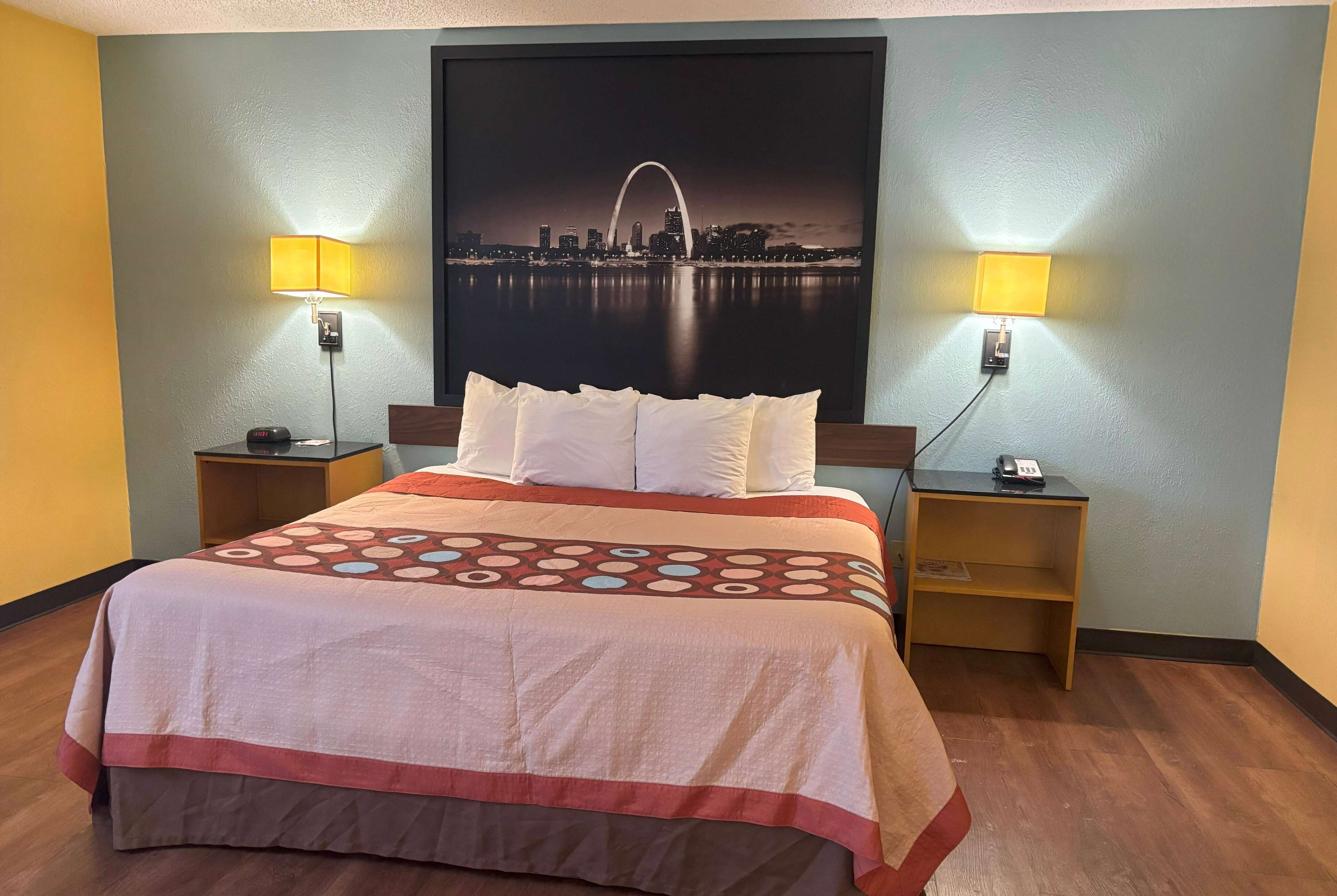 Quarto de hotel com cama, duas luminárias de cabeceira, mesa de apoio, telefone fixado na parede e pôster de paisagem urbana.