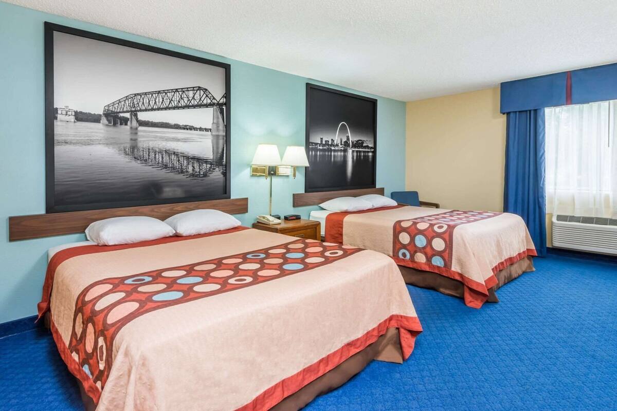 A hotel room with two beds, a large window with curtains, a wall-mounted air conditioner, and two wall-mounted lamps.