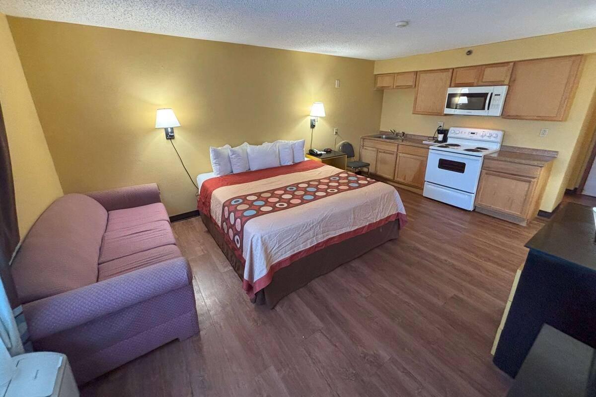 A compact hotel room with a bed, a sofa, a kitchenette with a microwave and stove, and two wall-mounted lamps.