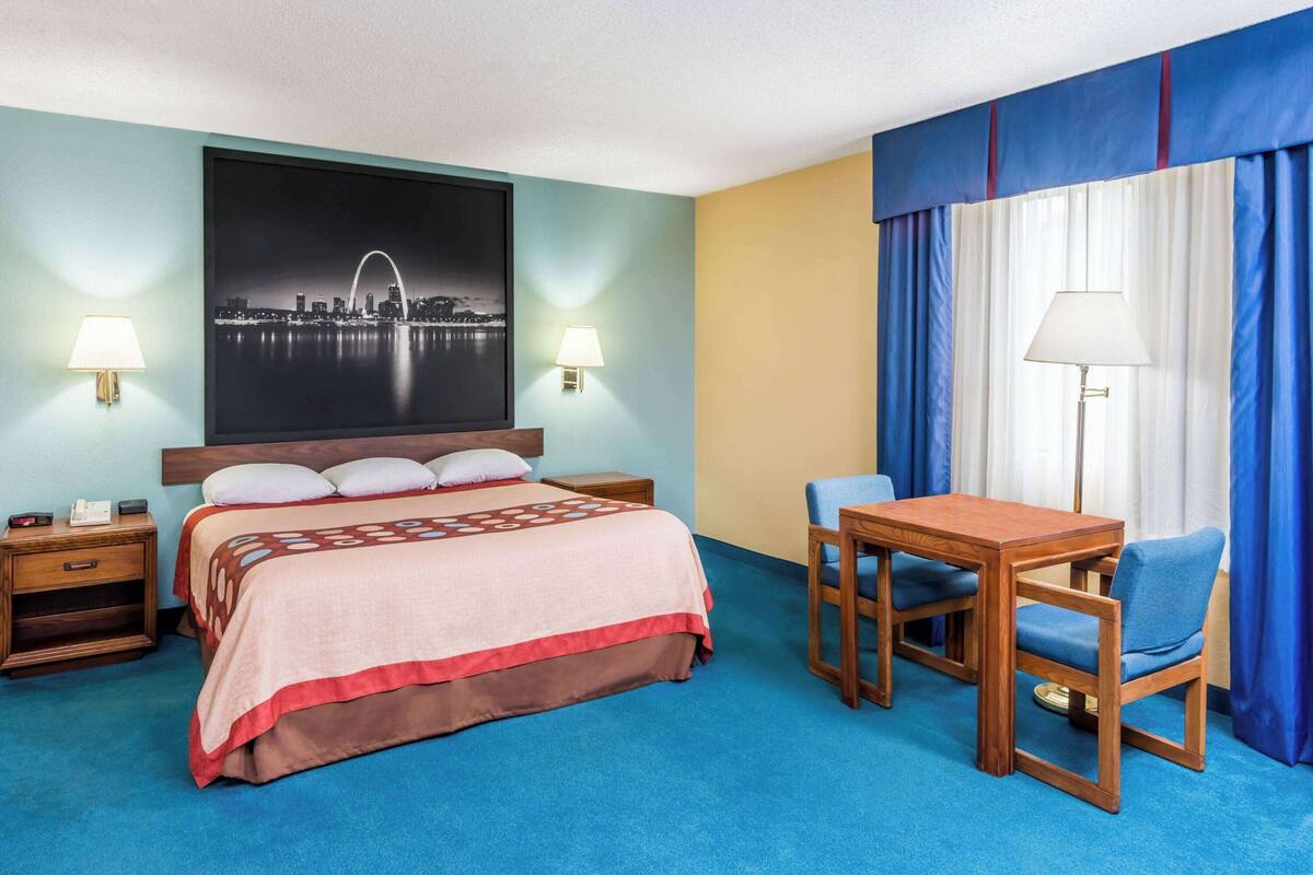 A hotel room with a bed, two bedside tables, a desk with a chair, a large framed picture on the wall, and a window with curtains.