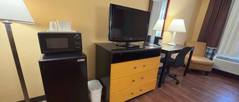 A hotel room with a desk, a television, a microwave, a small trash bin, and a chair.